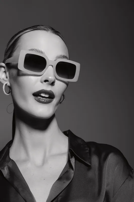 Stylish Caucasian woman with pale skin wears large sunglasses and silk blouse in studio. Fashion concept with confident female model in black and white.