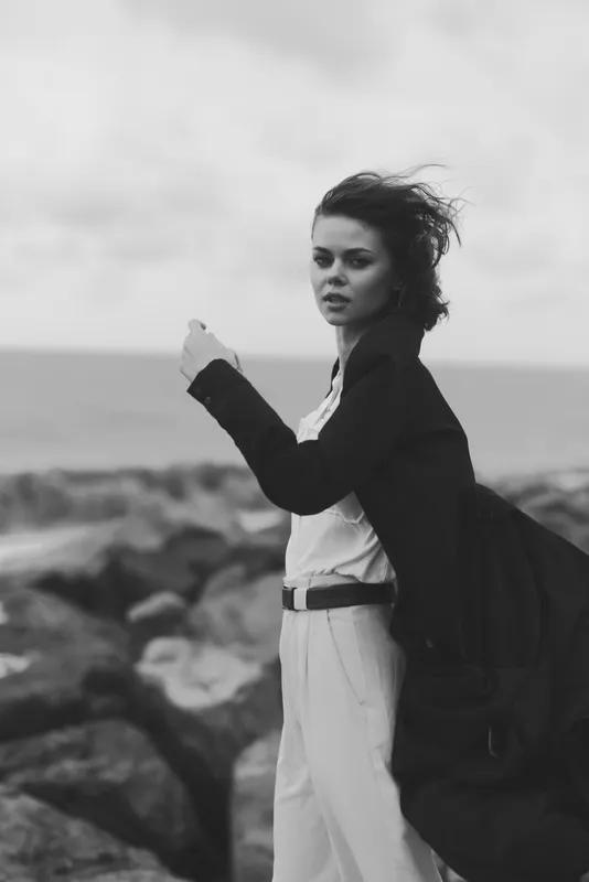 Woman on a rocky coastline with wind in her hair, wearing a coat and backpack, monochrome seascape conveying moody atmosphere and ocean breeze
