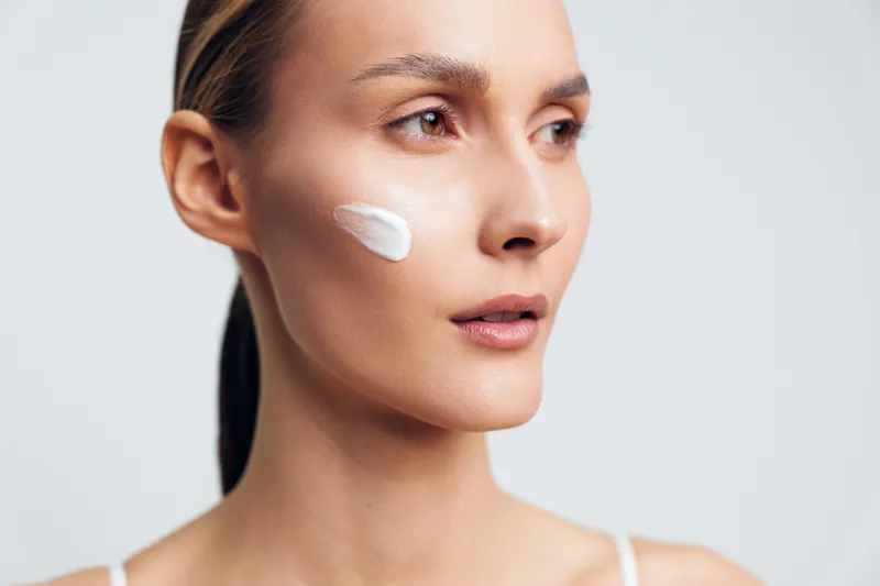 Young woman with cream on cheek looking sideways on light background. Concept of skincare, beauty, health, and self-care with soft natural lighting.