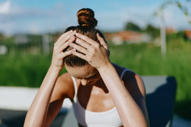 Woman sits outdoors with hands on her head, showing fatigue after a workout. Sunlit scene, casual athletic top, green lawn in background, conveys wellness, stress relief, and self care vibe.