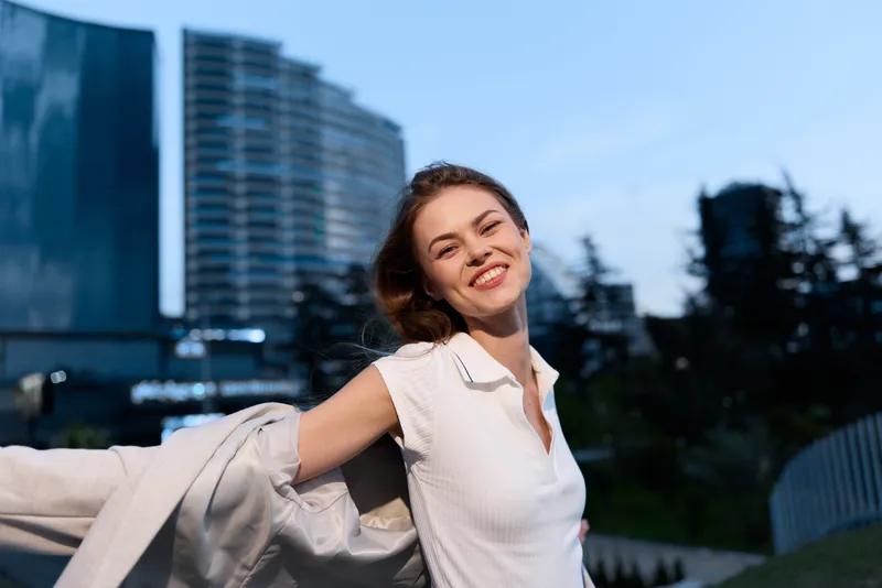 Confident woman in business attire smiling outdoors with urban background and soft evening light, showcasing modern professional style and positive energy.