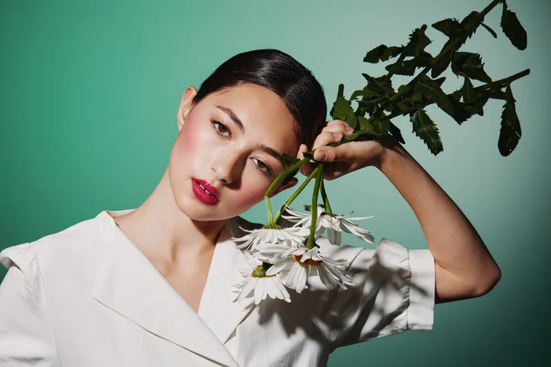 Stylish woman with vibrant red lips and sleek black hair holds a bouquet of white daisies against a green background. Creative beauty and fashion concept with high contrast lighting.