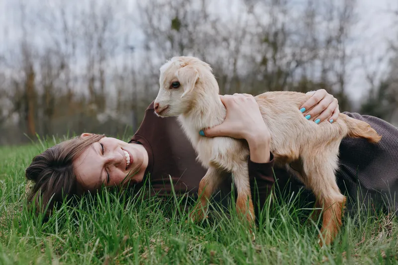 Smiling woman lying on grass in a rural field, embracing a goat kid. Warm natural light highlights gentle bonding, lifestyle moment, animal companionship, and outdoor wellness.