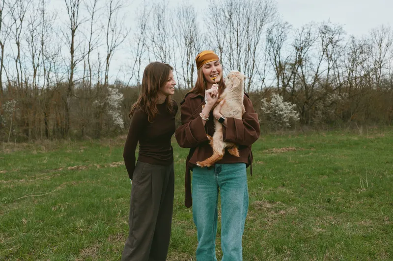 Cheerful women outdoors, smiling and sharing a moment with a goat kid, casual lifestyle in a grassy field, soft natural light, relaxed interaction, rustic vibe, natural colors, open countryside