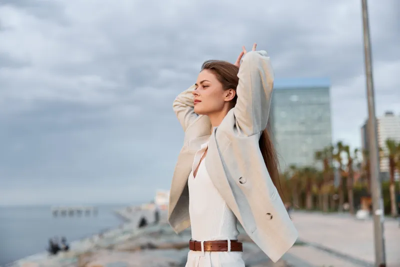 Woman in light beige coat outdoors on cloudy day with calm confident expression near urban waterfront, casual autumn fashion, serene mood, soft natural tones