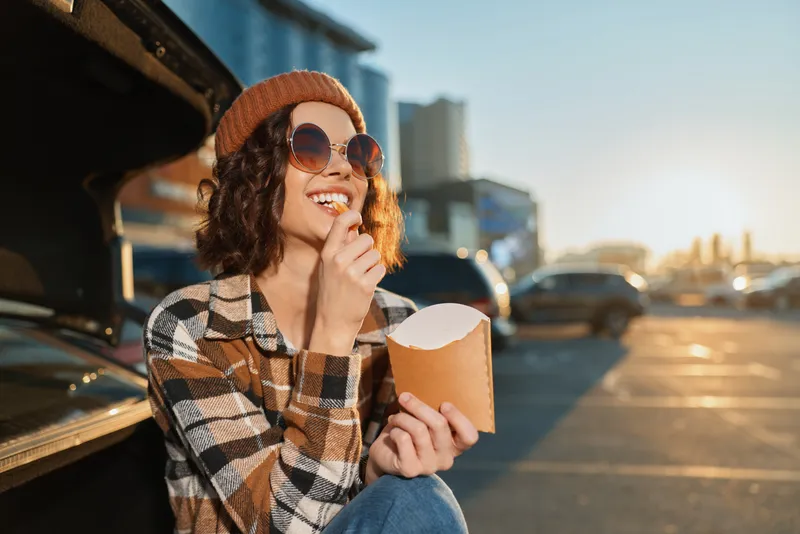 Woman eating fries by car trunk in parking lot at sunset, wearing sunglasses and beanie, smile and relaxed posture capture candid lifestyle and authenticity with golden hour glow and mindful living.