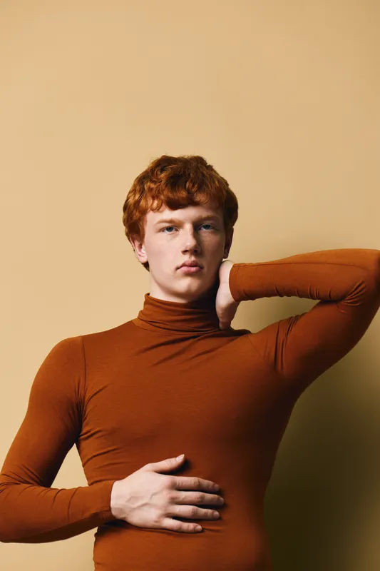 Young man with red hair and fair skin wearing orange turtleneck posing with hand on neck and stomach against beige background in studio portrait.