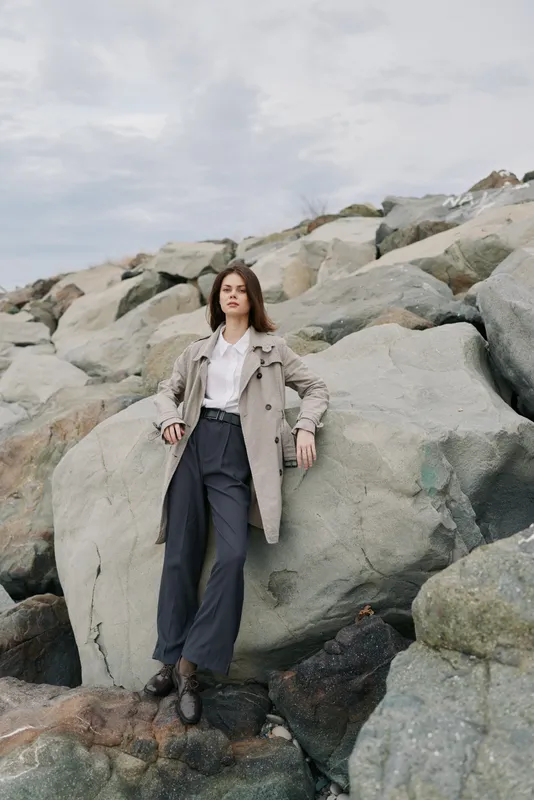 Young woman in stylish coat and black pants poses on large rocks outdoors under cloudy sky. Fashion portrait with natural landscape, casual outfit and confident model concept in nature.