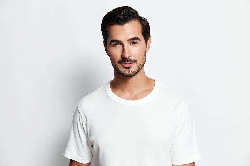 Confident young man standing against a plain white background, wearing a casual white t-shirt, looking directly at the camera with a charming smile. Fashion lifestyle concept.