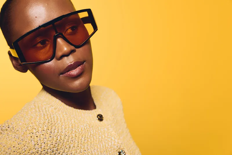 Portrait of a confident black woman wearing oversized sunglasses and a beige sweater against a yellow background. Concept of fashion, style, and modern beauty for dark skin models.
