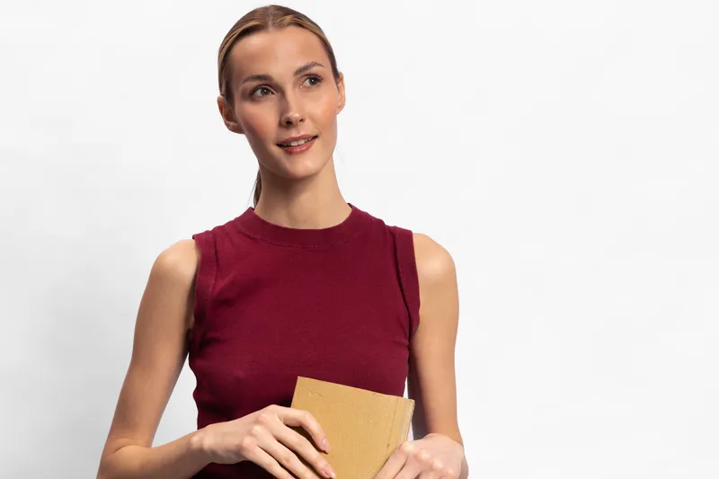 Young woman in sleeveless red top holds a wrapped gift box and looks away. Studio portrait on white background with soft lighting. Concept of celebration, surprise, and thoughtful gesture.