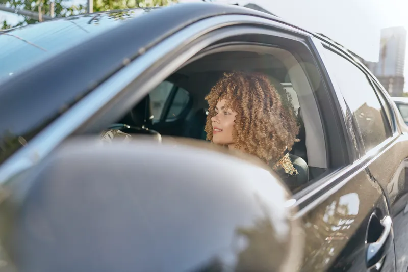 young woman driving car with curly hair smiles enjoying sunny urban day in modern black vehicle interior and exterior, casual lifestyle and positive mood