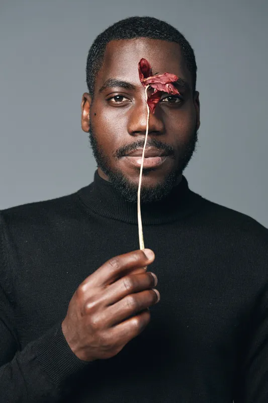Creative conceptual portrait of African man holding dried flower in front of face with serious expression, visual style with dark skin and black turtleneck on gray background.