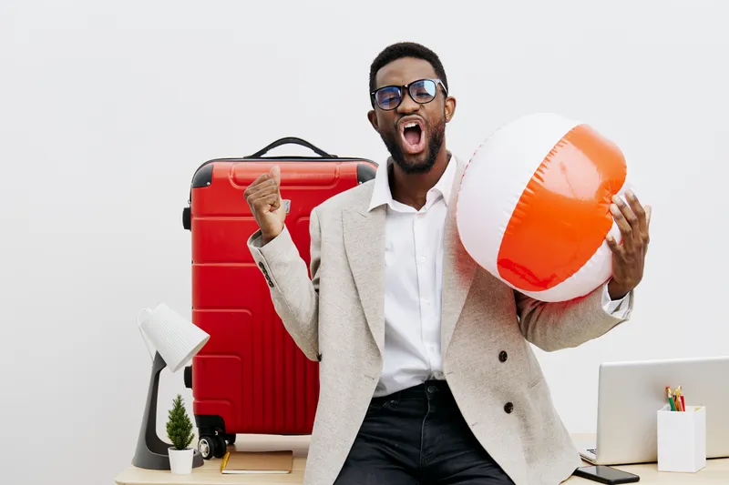 Happy excited young man with sunglasses holding a beach ball and celebrating in a cheerful office setting with travel accessories, red suitcase, laptop, and desk decor. People lifestyle concept for va