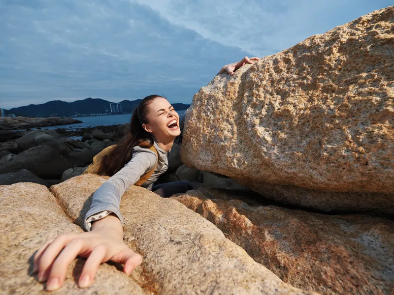A young female model exclaims joy while playfully interacting with large rocks at a coastal location, showcasing her excitement amidst a serene background