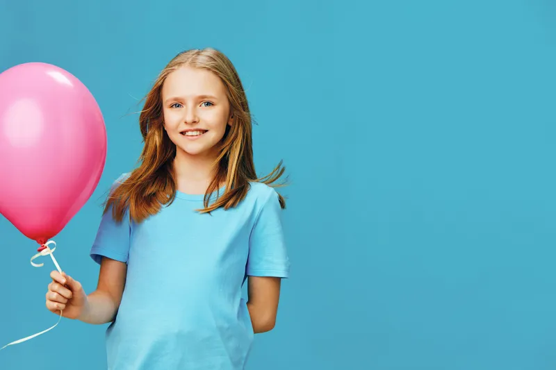 Smiling girl with a pink balloon against a blue background, celebrating joy and happiness, dressed in a casual light blue t shirt, perfect for seasonal events like birthdays