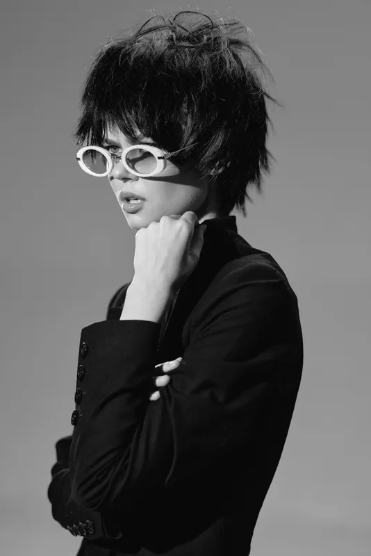 Black and white portrait of a stylish woman wearing white sunglasses and a black suit with thoughtful expression.