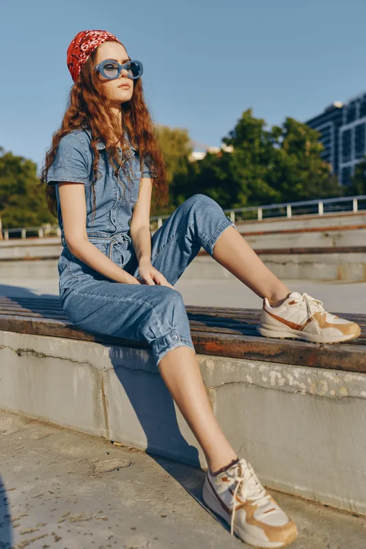 young woman wearing denim jumpsuit and sneakers sitting outdoors on concrete steps with sunglasses and red headscarf on sunny day