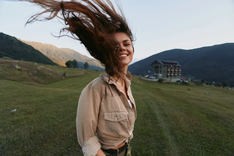 A smiling woman stands in a grassy landscape with distant mountains, wind in her hair, wearing a casual beige shirt. An outdoor lifestyle portrait captured in natural light.