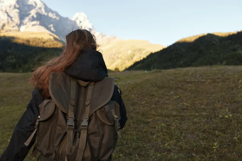 A lone hiker with a large backpack stands facing a vast mountain landscape. Distant peaks rise above rolling valleys, while clear skies and rugged terrain evoke adventure and exploration.