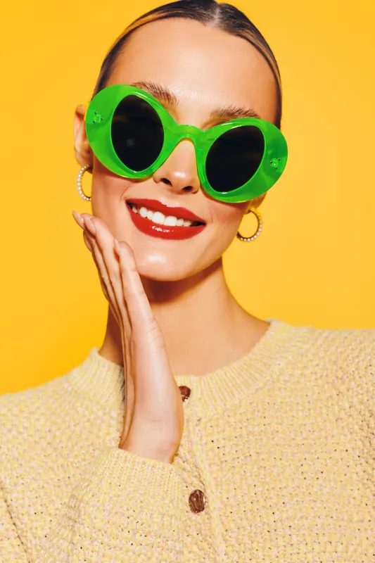 Smiling woman with bright red lipstick wearing large green sunglasses and yellow sweater poses against vibrant yellow background. Concept of fashion, summer style, and joyful mood.