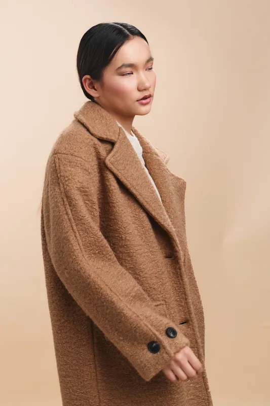 asian woman in a warm tan coat stands against a soft backdrop, exuding calm confidence and poised professionalism in a clean, modern mood that emphasizes style and composure.