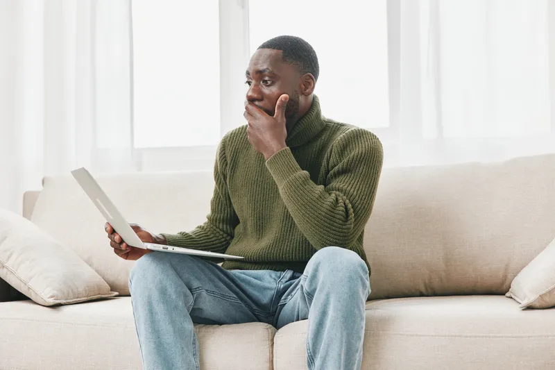 African man experiencing stress while working from home, wearing a green sweater, looking worried while using a laptop, in a bright living room