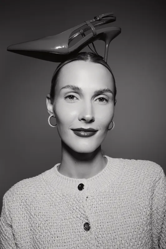 White woman with fair skin wears shiny high heel shoe on head, posing in textured sweater and hoop earrings on grey background.