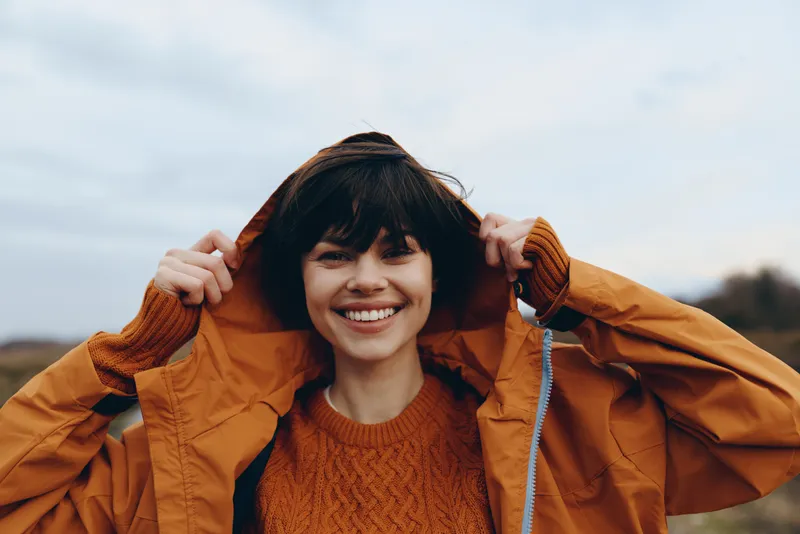 Woman outdoors in autumn, smiling portrait, wearing jacket with hood, casual lifestyle fashion. Young brunette holds hood with hands, relaxed happiness in cloudy daylight park.