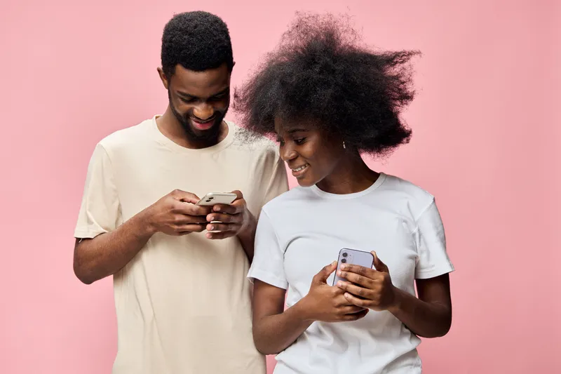 Happy young couple interacting with smartphones, dressed in casual clothing, set against a soft pink background, showcasing modern lifestyle and technology