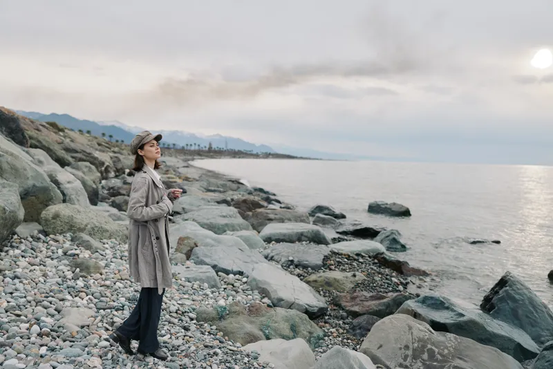 Woman in coat and hat stands on rocky beach looking at calm sea under cloudy sky. Concept of solitude, nature, and peaceful outdoor moment by waterfront.