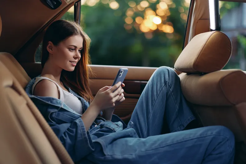 Relaxed girl in a denim outfit using a smartphone in the backseat of a car during sunset, showcasing a casual lifestyle with natural lighting.