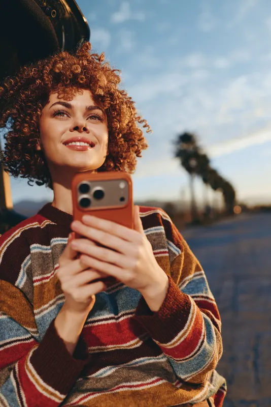 Woman lifestyle outdoors, curly haired woman with smile for selfie, holding smartphone near face, sunset evening light, street background blurred, casual sweater, relaxed portrait