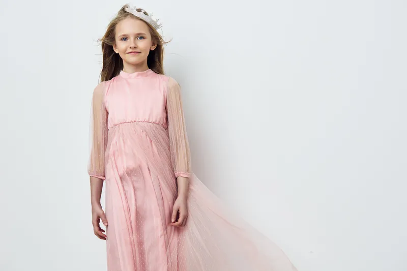 Beautiful girl in a flowing pink dress with sheer sleeves stands gracefully against a soft white background, exuding innocence and joy Perfect for children s fashion photography