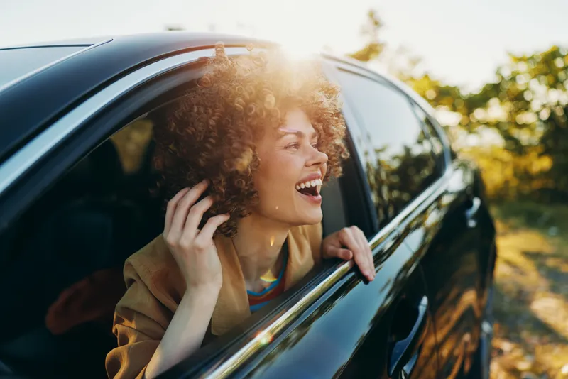 A joyful woman leans out of a car window, enjoying warm sunlight, capturing a candid travel moment with retro film color and carefree mood, featuring natural lighting and a sense of freedom.