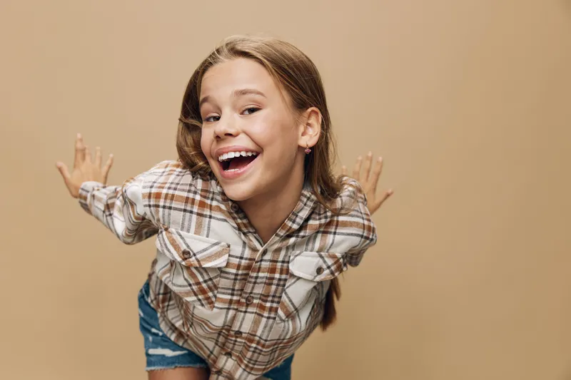 Girl smiling and posing, wearing checkered shirt and denim shorts on beige background. Happy child expressing joy, playful and energetic mood in casual outfit with long hair.