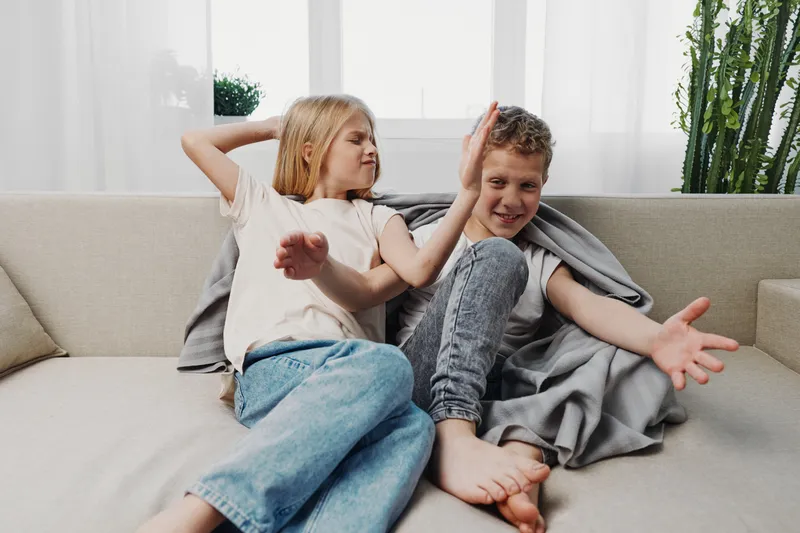 Two children, a boy and a girl, playfully interact on a beige couch in a bright living room with natural light and indoor plants. Concept of sibling fun, childhood and family moments.