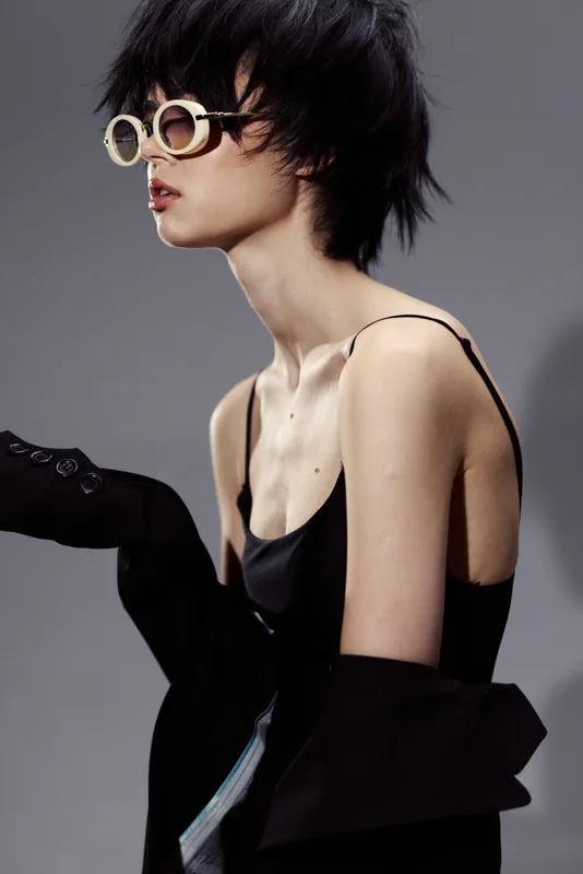 Stylish woman in sunglasses wearing a black dress with an off-shoulder jacket posing on a gray background