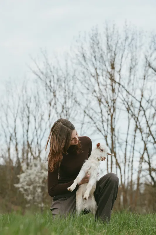 Woman sits on grass in a park, embracing a fluffy white dog, sharing affection and calm companionship, with bare trees and soft natural light creating a tender lifestyle moment in spring air.