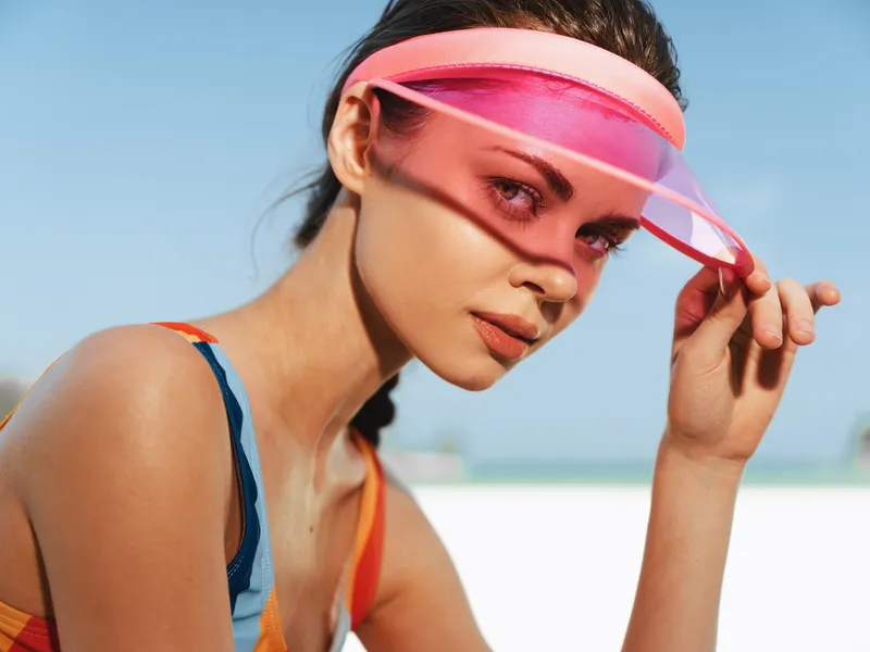 Close up portrait of a stylish woman at the beach wearing a pink translucent visor, sunlit skin, and colorful swimwear, capturing vibrant summer fashion and confidence.