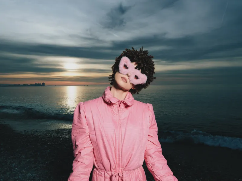 Beach scene with a woman in a pink robe wearing a soft decorative mask during sunset, relaxed pose by the shore, cinematic glow, serene mood and playful style.