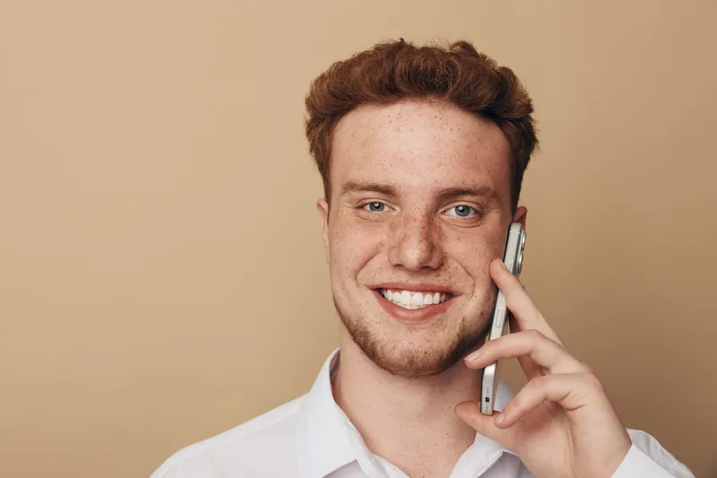 Smiling young man with red hair and freckles talks on smartphone against beige background. Casual portrait of happy male using mobile phone for communication and social connection.