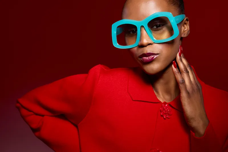 Stylish African woman with dark skin wearing bright blue oversized glasses and red outfit posing confidently against red background. Fashion portrait with bold colors and modern accessories.