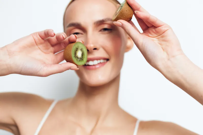 Cheerful woman playfully holds kiwi and fruit halves to her eyes, bright portrait mood, refreshing natural glow, healthy skin, relaxed pose, light background, casual style, joyful moment across frame