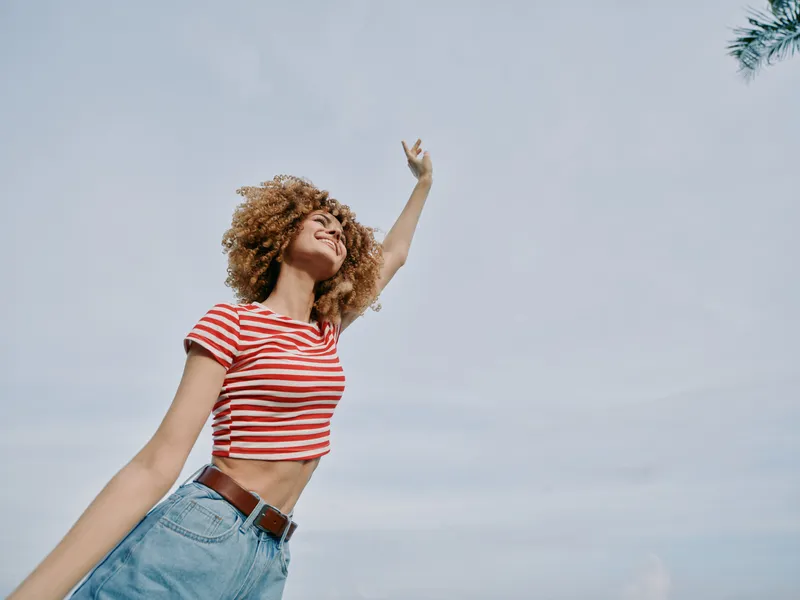 A joyful young woman with curly hair wears a red and white striped crop top and light denim jeans, posing outdoors with arms raised toward a bright blue sky.