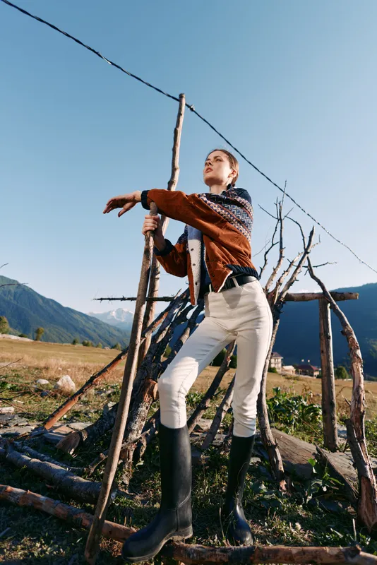 Woman outdoors in mountains leaning on a wooden structure in a rural field, wearing textured sweater, white pants and black boots, portrait with clear sky, sunlight and rustic mood.