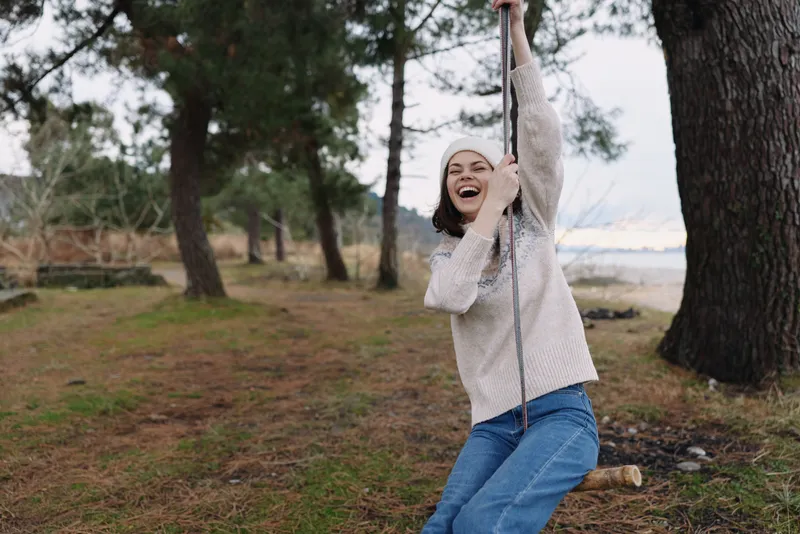 Woman enjoying a swing outdoors in a park, laughter and carefree moment among trees and grass, rope swing in motion near a calm shoreline and gentle breeze