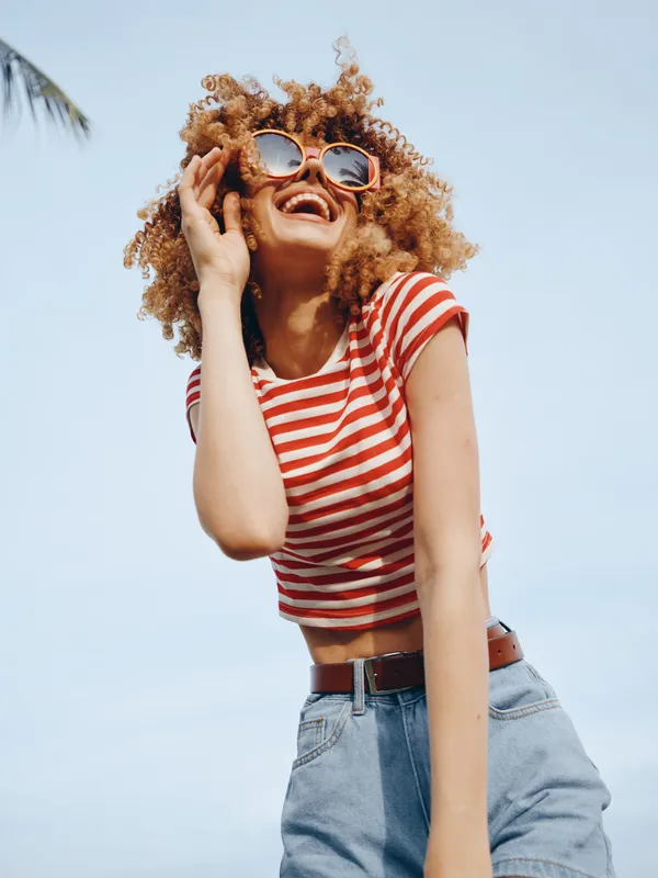 Vibrant portrait of a young woman with curly hair, wearing a red and white striped tee and jeans, sunglasses, posing outdoors on a sunlit summer day against a clear blue sky.
