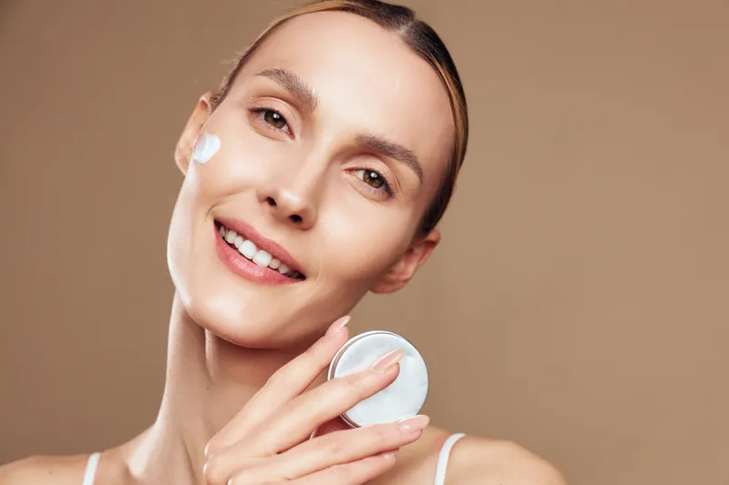 A smiling woman applies moisturizing cream to her face with a cotton pad against a beige background. The concept focuses on skincare, beauty routine, and healthy skin care.