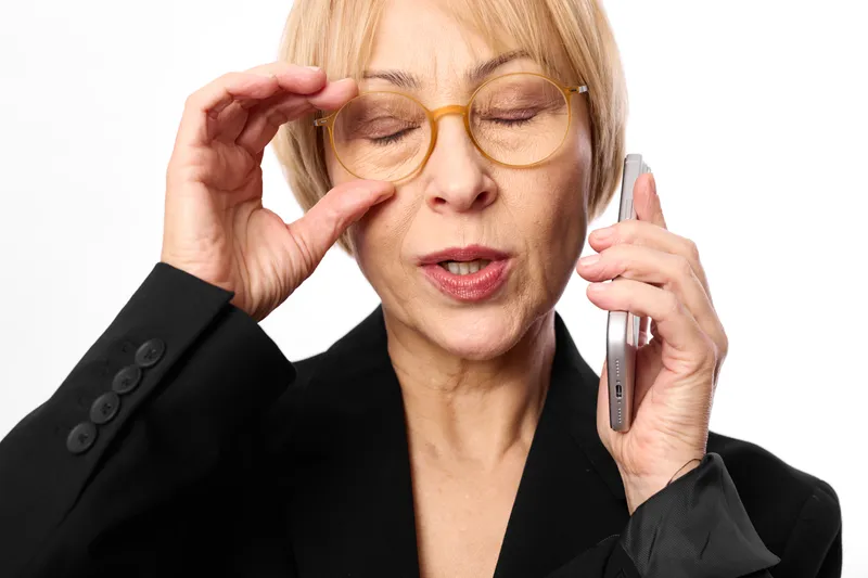 Stressed older business woman holding a phone to her ear, adjusting glasses and rubbing her forehead, tired face on white background, office pressure concept.